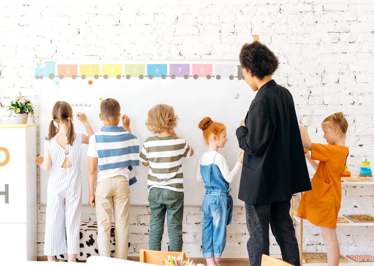 Group of children and teacher interacting with a whiteboard in a bright, indoor classroom setting.